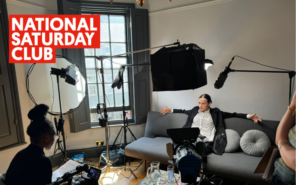 Image of a young person on a sofa on a film set. There is another person sitting far left directing her