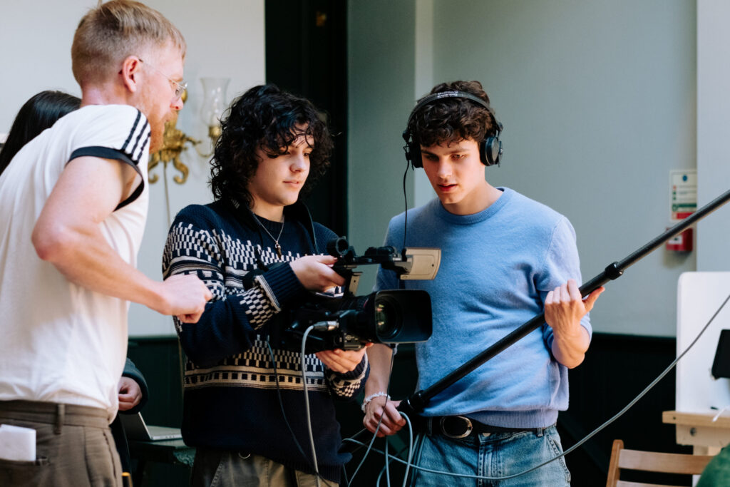 Two young men holding film equipment overseen by tutor in white t-shirt
